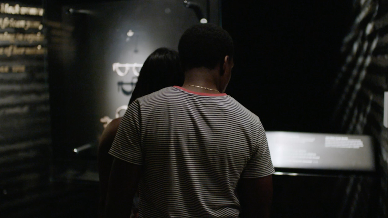 An individual who can be read male hugs another individual from behind while they both look at an exhibition display in the National Museum of African American History and Culture. Film still by Oliver Hardt